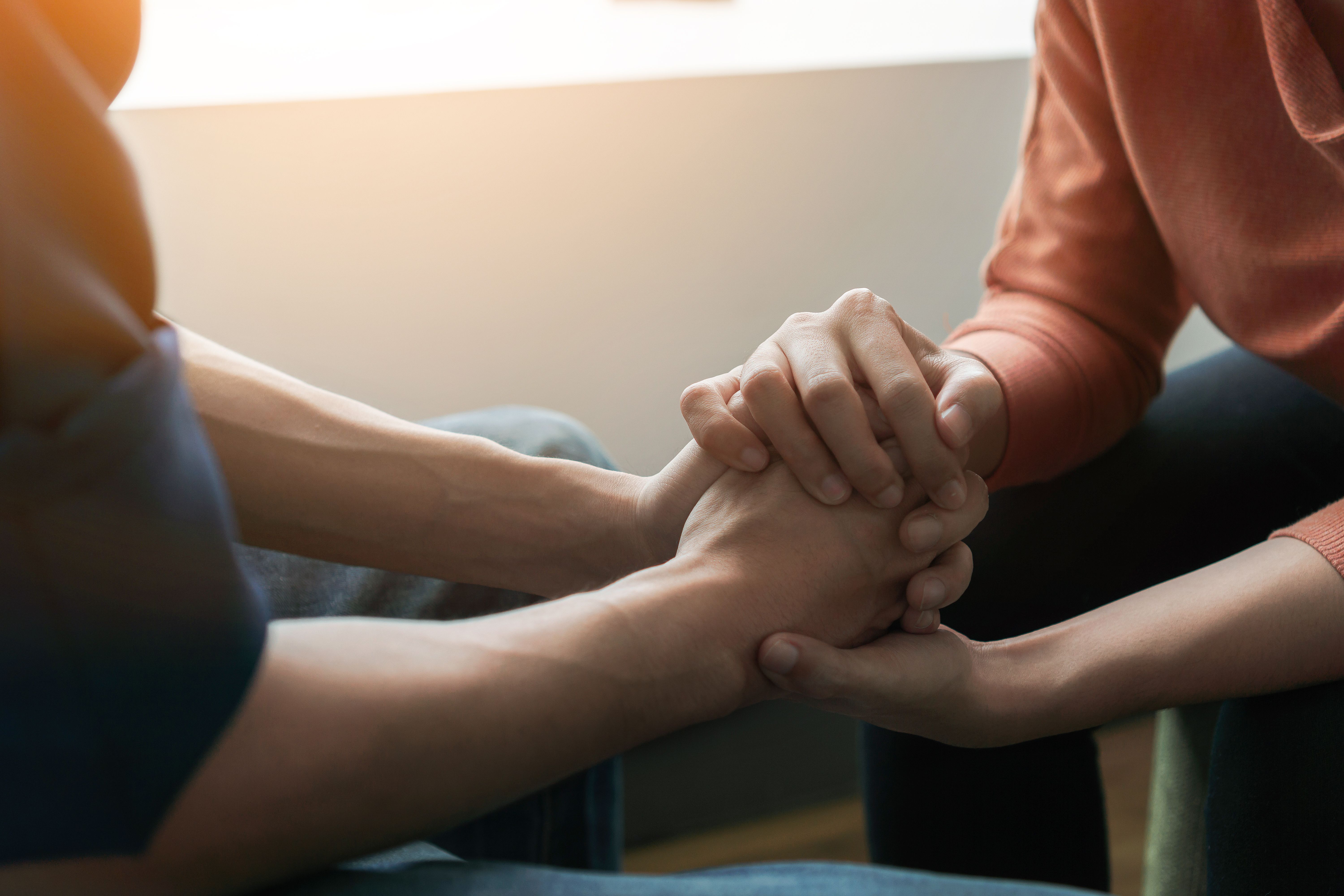 Dwie osoby siedzące blisko siebie, trzymające się za ręce w pocieszającym i wspierającym geście, z miękkim naturalnym światłem oświetlającym scenę.